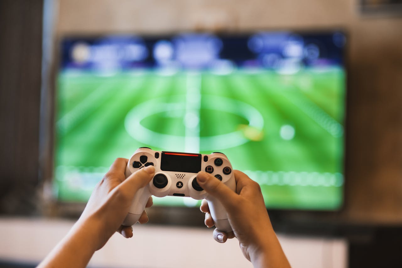 about-img Close-up of hands holding a game controller playing a football video game on screen.